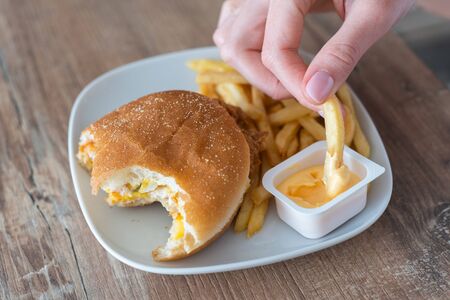 Harmful cheeseburger and fries. The hand dips the fried potatoes into the sauce. Fast food on a...