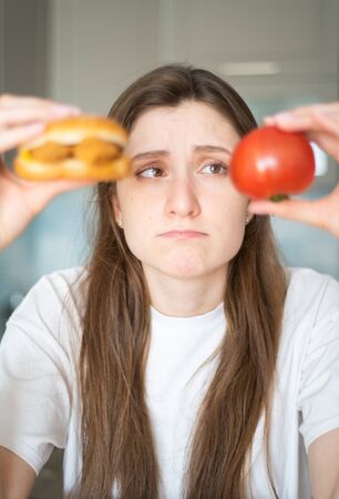Temptation of junk food. The girl is thinking about what to eat. A beautiful woman holds a tomato and cheeseburger and looks at themの写真素材
