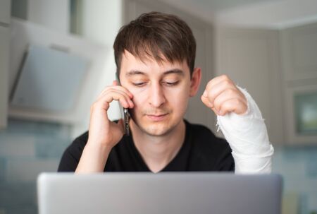 Man with fracture arm communicates on the phone behind a laptop. Problems with insuranceの写真素材