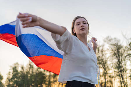 The face of a beautiful woman with a Russian flag in her hands. Pretty woman walks with a flag behind her back against the sunset skyの写真素材