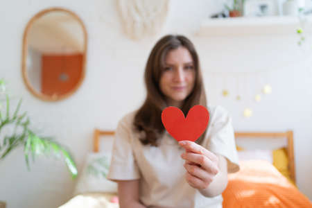 Give love on Valentines Day. Red paper heart in the hand of a young woman in a warm room