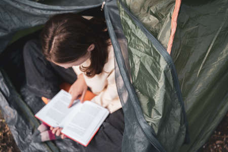 Reading a book in a tent. Relaxing holidays in nature. A young woman rests in a tent campの写真素材