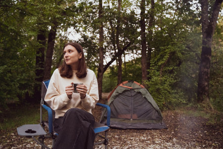 Woman backpacker drinks from a hot mug in the forest. Tent in the campsite. Adventure, discovery of...