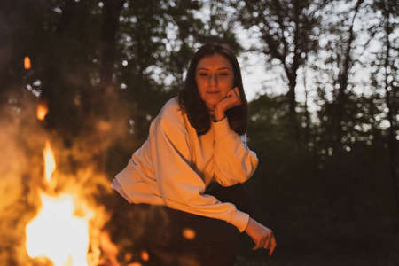A young woman sits next to a fire in a camp. Warm and cozy campfire