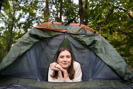 Portrait of a tourist woman peeking out of a green tent in the woods in nature. Adventure, journey...