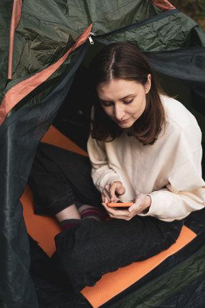 A young tourist sits in a tent in nature and looks at the phone. Surfing the Internet outside the city on hikeの写真素材