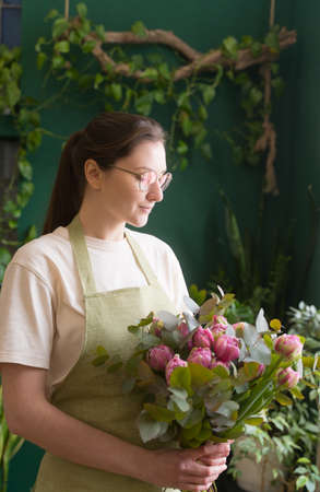 Businesswoman. Young woman florist in an apron and glasses checks a floral bouquet. Beautiful flowers to order. The concept of young entrepreneursの写真素材