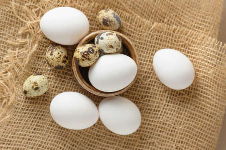 White raw chicken eggs and small quail eggs in a clay bowl on a burlap close-up. Chicken and quail eggs rustic background.の写真素材