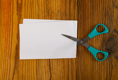 Scissors cutting folded paper. Blue scissors cuts white paper piece on wooden background top view