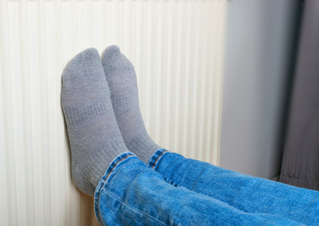 Warming Feet on White Radiator, Man Warms his Feet at Home, Cold Winter, Expensive Electricity Saving Concept, Copy Spaceの写真素材