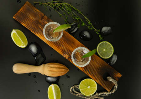 Two stacks of tequila with fresh lime slices and salt on stylish wooden board isolated on black backgroundの写真素材