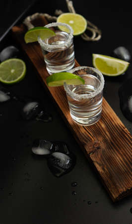 Two stacks of tequila with fresh lime slices and salt on stylish wooden board isolated on black backgroundの写真素材