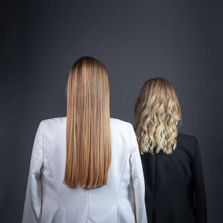 two girls with long blond hair stand against wall in strict suits, rear viewの写真素材