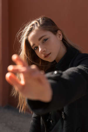 Young beautiful girl in black coat on sunny day stretches her hand forward. Hand out of focus....
