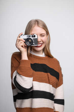 Young pretty blonde girl with bright red lips smiling, holding vintage photo camera, taking picture on white backgroundの写真素材