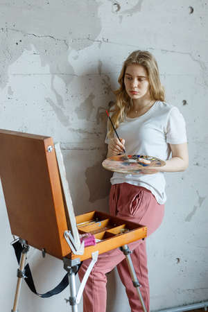 stay home: young girl with brush and palette standing near easel drawing pictureの写真素材