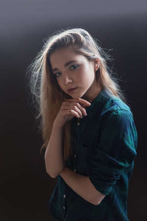 young pretty girl in checkered shirt and backlit hair poses on black background, tilts her head and touches her chinの写真素材