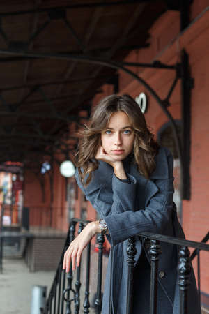 young thoughtful girl in gray coat and turtleneck poses in front of red buildingの写真素材