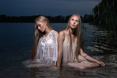 two young twin sisters posing in light dresses in water of lake at summer nightの写真素材