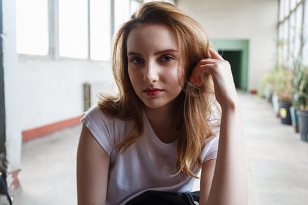 young caucasian girl sits in t-shirt at coridor with windows and houseplants