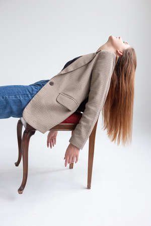 portrait of young caucasian woman with long hair in black top, blue jeans and suit jacket, lying on chair at studio with her chin up, throwing her head back. pretty lady posing on white backgroundの写真素材