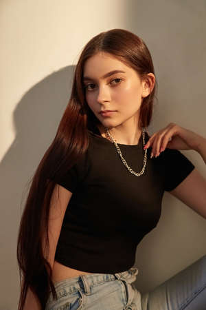 portrait of young caucasian lady with long hair in black top and blue jeans sitting next to wall....