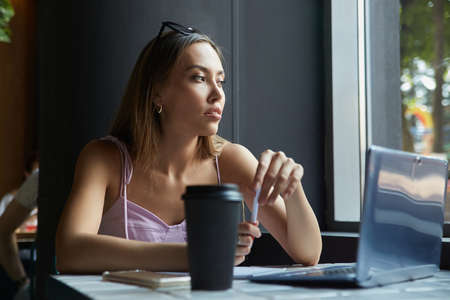 beautiful young asian woman sitting at table near window, working on laptop. beautiful lady...