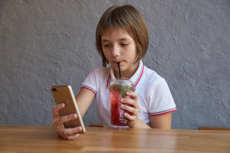 little schoolgirl holds smartphone, drinks lemonade through straw. girl reading information on phone, drinking red icy cocktail at table of cafe. modern communication technology, using digital devicesの写真素材