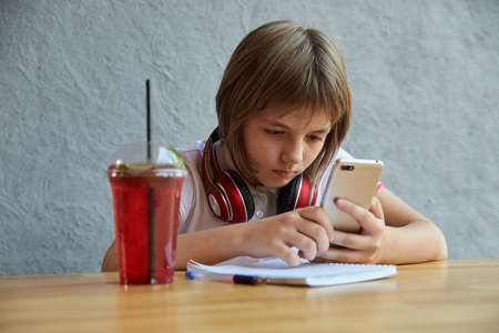 schoolgirl with headphones and red icy lemonade holds smartphone in hands. little girl using mobile phone at cafe. modern communication technology, using digital devices, gadgets, social networksの写真素材