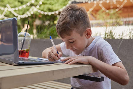 little schoolboy doing homework outdoors with laptop. smiling caucasian boy sitting at table at veranda of cafe, studying with computer, writing notes in copybook. distant learning, online educationの写真素材