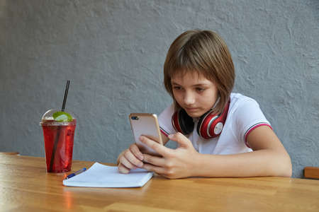 schoolgirl with headphones and red icy lemonade holds smartphone in hands. little girl using mobile phone at cafe. modern communication technology, using digital devices, gadgets, social networksの写真素材