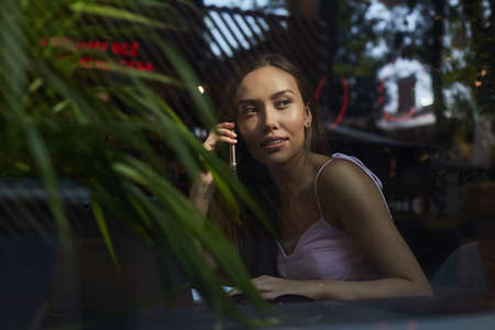 young asian pretty woman sitting at table of coffee shop, calling by smartphone. beautiful lady using cell phone shot through window glass outside of cafe. modern communication technologyの写真素材
