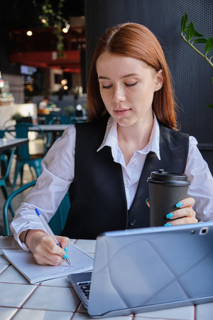 beautiful young caucasian business woman sitting indoors, writing notes in notebook. pretty student sitting at table of cafe, studying with laptop, doing homework. distant work, online educationの写真素材