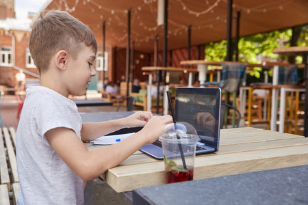 little schoolboy sits outdoors, types on keyboard of laptop. smiling caucasian boy sitting at table at veranda of cafe, studying with computer, doing homework. distant learning, online educationの写真素材