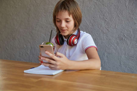 schoolgirl with headphones and red icy lemonade holds smartphone in hands. little girl using mobile phone at cafe. modern communication technology, using digital devices, gadgets, social networksの写真素材