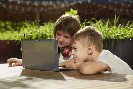 boy and girl sit outdoors, watch at screen of laptop on sunny day. little caucasian schoolchildren...