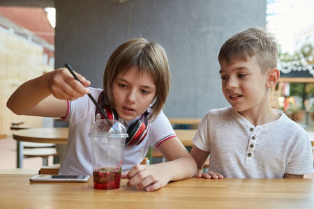 boy and girl sit outdoors at veranda of cafe. little caucasian schoolchildren sitting at table, playing with berries in cup of lemonade. childhood concept, fun, joyの写真素材