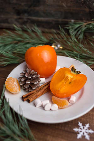 Christmas still life with fresh persimmon on a plate with pine cones, cinnamon, marshmallow and...