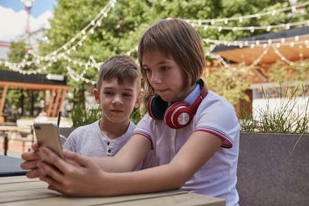 children sit outdoors, watch at screen of smartphone. little caucasian schoolchildren sitting at table at cafe veranda, using mobile phone. modern technology, gadgets, digital devices, entertainmentの写真素材