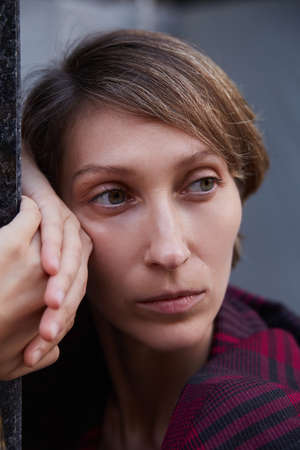 melancholic lonely young woman with short hair in red checkered jacket outdoors in front of building. closeup portrait of caucasian female leaning on wall with her arms and headの写真素材