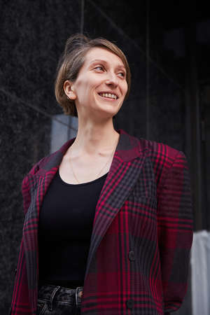 young woman with short hair in red checkered jacket outdoors in front of building. portrait of...