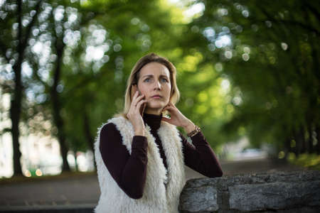 Beautiful young woman in summer. Blonde girl is talking on the cell phone outside in city nature. Female in a white furry jacket, brown sweater and with telephone. Businesswoman calls with smartphoneの写真素材