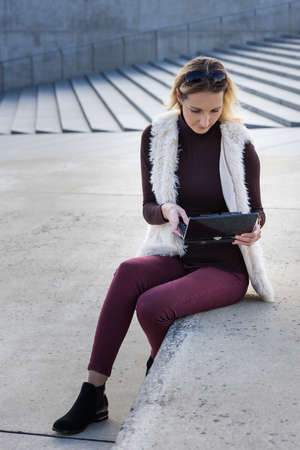 Beautiful young woman close up. Blonde girl is sitting on steps outside an office, holding a tablet. Female in a white furry jacket, red shorts, with notebook. Businesswoman doing the computer work.の写真素材