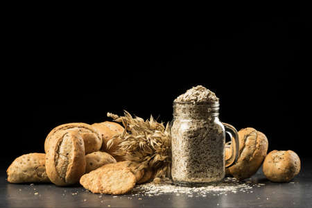 Oat bunch, baked white bread, cookies and flakes in flavouring jar, isolated on black background. Grain bouquet, golden oats spikelets on dark wooden table, buns and can filled with dried grains. Food, bakery conceptの写真素材