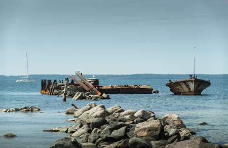 Old broken boat wreck on the shore, blue sea and sailboat on background. Harilaid, small island in Estonia, Europe.の写真素材