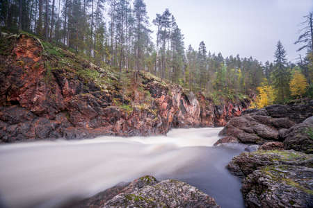 Red cliff, stone wall, forest, waterfall and wild river view in autumn. Fall colors - ruska time in KiutakÃ¶ngÃ¤s. One part of Karhunkierros Trail. Oulanka National Park, north Finland, Lapland, Europeの写真素材