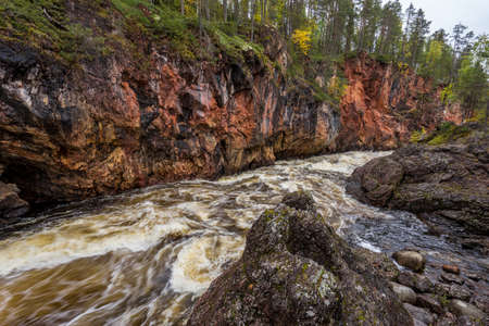 Red cliff, stone wall, forest, waterfall and wild river view in autumn. Fall colors - ruska time in KiutakÃ¶ngÃ¤s. One part of Karhunkierros Trail. Oulanka National Park, north Finland, Lapland, Europeの写真素材