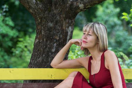 Young blond woman in a red dress leaning sits on a wooden bench.