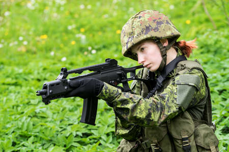 Portrait of armed woman with camouflage. Young female soldier observe with firearm. Child soldier with gun in war, green goutweed background.  Military, army people conceptの写真素材