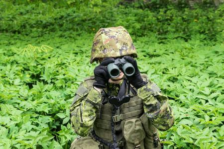 Portrait of armed woman with camouflage. Young female soldier observe with binoculars. Child soldier with gun in war, green goutweed background.  Military, army people conceptの写真素材